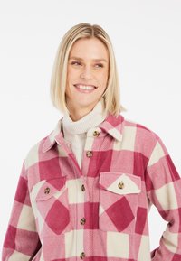 Chemise à carreaux rose et crème, fabriquée en tissu doux. Comprend deux poches poitrine et un col arrondi. Fermeture à boutons à l'avant.