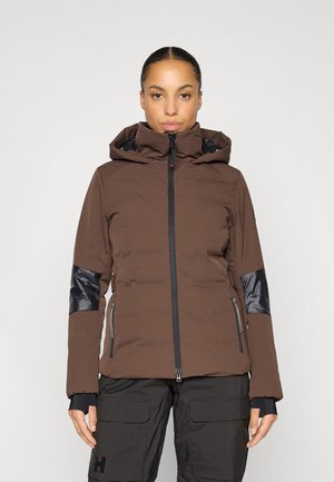 Frau trägt eine braune Kapuzenjacke mit schwarzen Ellbogenflicken und schwarzen Reißverschlusstaschen, kombiniert mit schwarzen Cargohosen.