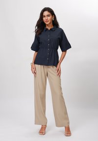 Top blu navy con bottoni, vita arricciata e maniche ampie, abbinato a pantaloni larghi di un beige chiaro e sandali con cinturini. Tessuto dalla superficie liscia.