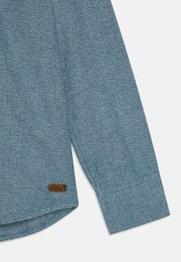 Blå skjorte med fiskebeinsmønster på ermet, med en teksturert overflate, og en lærdetalj nær mansjetten. Mykt stoff, skreddersydd design.