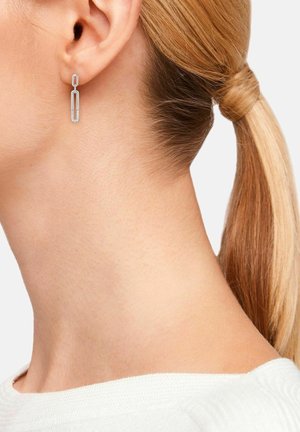 Femme aux cheveux blonds attachés en queue de cheval portant une boucle d’oreille en chaîne argentée avec de petites pierres incrustées sur le maillon inférieur.