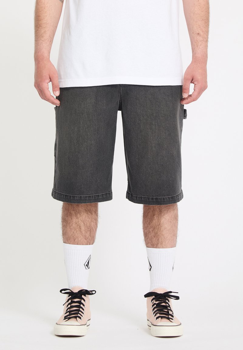 Homme portant un short en denim noir jusqu'aux genoux, des chaussettes blanches montantes avec des logos noirs, et des baskets roses à lacets noirs, debout sur un fond blanc.