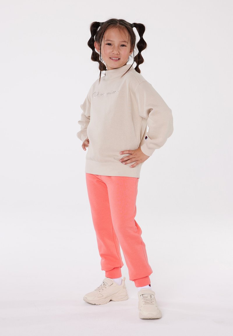 Beige long-sleeve sweatshirt with embroidered text, paired with coral sweatpants and white sneakers, displayed against a white background.