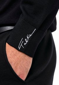 Cuff in maglia nera con logo "Falke" ricamato in bianco. Tessuto testurizzato con una vestibilità aderente, che mette in evidenza i dettagli nella cucitura.