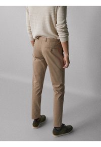 Pantaloni beige aderenti con dettagli sulle tasche posteriori, abbinati a scarpe verde scuro. Il tessuto appare liscio con un design su misura.