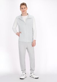 Helles grauer Reißverschluss-Weste über einem weißen Langarmshirt, kombiniert mit grauen Jogginghosen und weißen Sneakers. Glattes Material, lässige Passform.