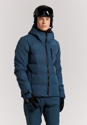 Joven con chaqueta de esquí azul, pantalones a juego, guantes negros y casco de esquí negro con gafas sobre un fondo liso.