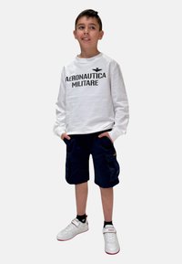 Camicia bianca a maniche lunghe con stampa "AERONAUTICA MILITARE", abbinata a pantaloni cargo blu scuro. Sneakers bianche con dettagli rossi.