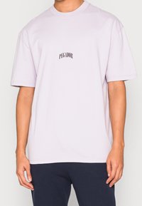 Muž v světlém purpurovém tričku s krátkým rukávem, na kterém je uprostřed hrudi černý nápis "PEGADOR", ve spojení s tmavými kalhotami.