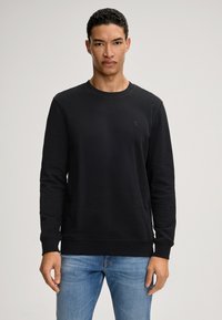 Schwarzer Rundhals-Sweatshirt mit langen Ärmeln, gerippten Bündchen und Saum. Mit dezentem Logo auf der Brust. Kombiniert mit hellblauen Jeans.