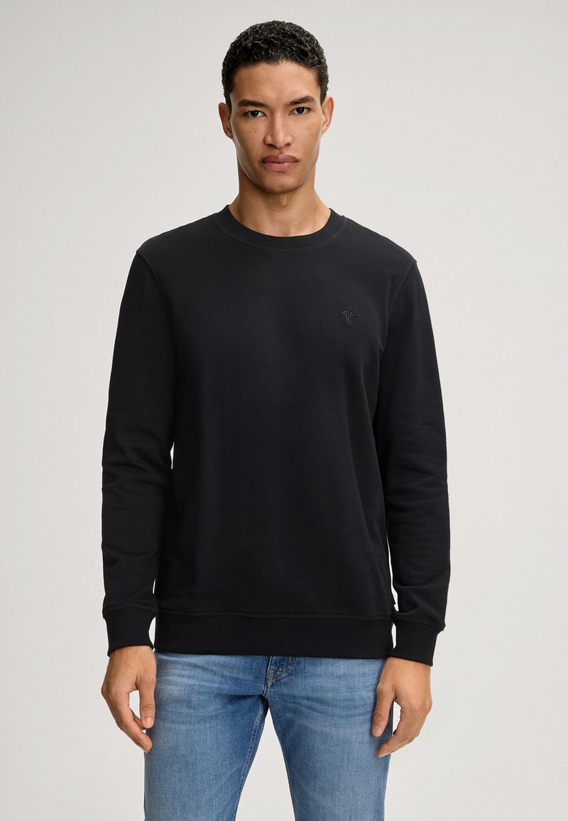 Schwarzer Rundhals-Sweatshirt mit langen Ärmeln, gerippten Bündchen und Saum. Mit dezentem Logo auf der Brust. Kombiniert mit hellblauen Jeans.