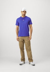 Camiseta de algodón morada con un pequeño logo blanco de Air Jordan, pantalones caqui y gorra blanca con letras negras. Zapatillas grises y blancas completan el conjunto.