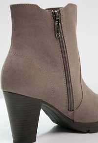 Grå mocka ankelstövel med dragkedja på sidan, avsmalnande klack och kurvdesign; har en texturerad yta och en mörk träklack som accent.