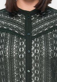 Blouse noire en dentelle avec des motifs floraux et géométriques complexes, boutonnée sur le devant, portée par une personne aux cheveux bruns et courts.