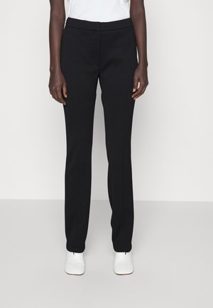 Person iført sorte bukser med lige ben og hvide sko, stående mod en ensfarvet lys baggrund.