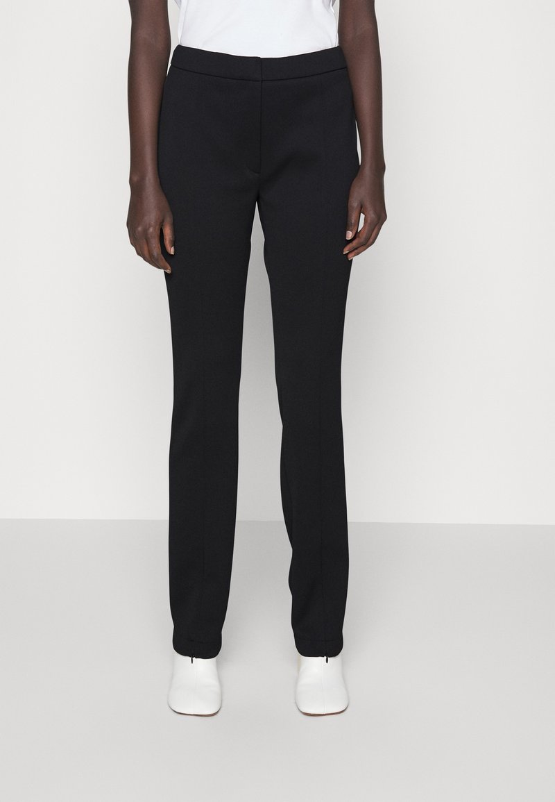 Personne portant un pantalon droit noir et des chaussures blanches, debout devant un fond clair uni.