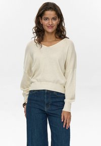 Hellbeige geripptes Sweatshirt mit V-Ausschnitt und weit geschnittenen Ärmeln, kombiniert mit dunkelblauen weiten Jeans. Weiche Textur, lässiger Schnitt, minimalistisches Design.