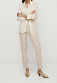 Jasnokremowa lniana koszula z guzikami, zestawiona z dopasowanymi kremowymi spodniami. Złote szpilki z ostro zakończonym noskiem i paski na kostkę, minimalne detale w całej stylizacji.