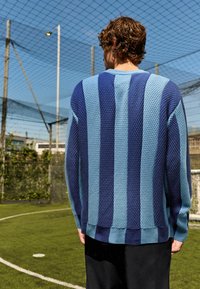 Gestreifter Pullover in Blau und Hellblau aus gestricktem Stoff mit strukturiertem Material, lockerer Passform und geripptem Saum. Getragen mit schwarzen Hosen auf einem Sportplatz.