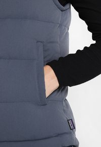 Grå puffervest med en synlig ficka, med en slät textur och en vävd etikett med en logotyp på nedre vänstra sidan.