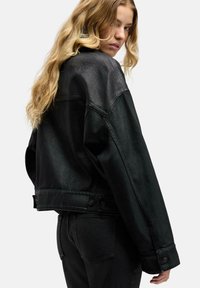 Jonge vrouw met lang blond haar, gekleed in een oversized zwarte spijkerjack en zwarte jeans, kijkend over haar schouder.