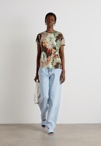 Brun skjorte med korte ermer og et flerfarget botanisk mønster, parret med lyseblå løse jeans og lyseblå sandaler. Modellen holder en hvit veske.