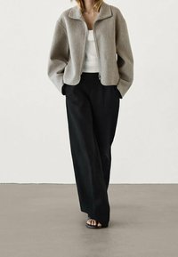 Jasnoszara kurtka typu cropped z zamkiem z przodu i szerokim kołnierzem, zestawiona z czarnymi spodniami o szerokich nogawkach i czarnymi sandałami na neutralnym tle.