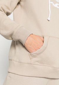 Ruka zasunutá v přední kapse béžové mikiny s ribbovanými manžetami a pasem, ukazující detail volného oblečení.