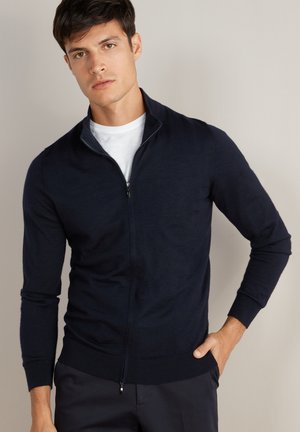 Giovane uomo in piedi con la mano in tasca, indossa un cardigan blu navy con zip sopra una T-shirt bianca, su sfondo beige uniforme.