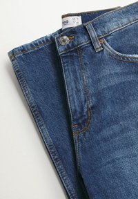 Jean en denim bleu plié avec taille visible, bouton, passants de ceinture et étiquette de marque indiquant la taille.