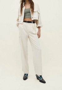 Witte wijde broek, beige cropped jasje met knoopdetails, olijfgroen cropped top en zwarte hakken met teal accenten.