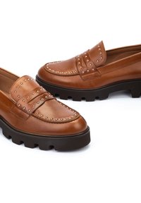 Mocasines de cuero marrón con acentos de tachuelas, puntera redondeada y suela negra gruesa, con superficies lisas y texturizadas.