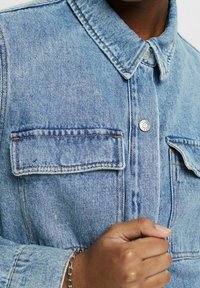 Ljusblå denimjacka med krage, två bröstfickor med lock och silverfärgade tryckknappsstängningar.