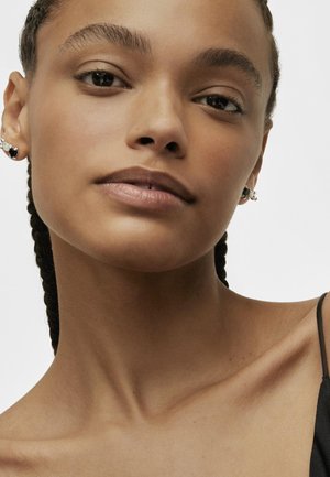 Primo piano di una persona con capelli intrecciati, che indossa orecchini d'argento con una pietra nera. Il tono della pelle è caldo e indossa un indumento nero.