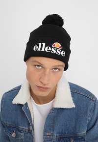 Black knit beanie with a pom-pom on top, featuring white embroidered "ellesse" text and a colorful logo. Worn with a denim jacket.