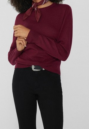 Femme portant un pull à manches longues bordeaux, un pantalon noir avec une boucle de ceinture argentée et un foulard bordeaux à motifs blancs.