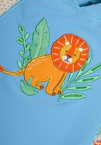 Vil�ágoskék rash guard, amelyen egy hímezett oroszlán látható, narancssárga arccal és sörénnyel, zöld levelek ölelésében, sima anyagon.