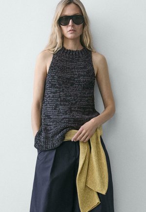 Donna che indossa un top nero senza maniche in maglia, pantaloni larghi scuri, grandi occhiali da sole neri e tiene in mano un maglione giallo in maglia contro un muro bianco.