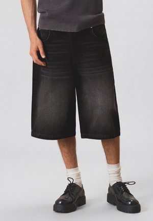 Mand iført sorte knælange denimshorts, hvide crew-sokker og sorte chunky snørebåndssko, står mod en ensfarvet baggrund.