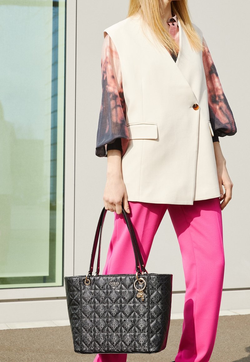 Personne portant un gilet crème, des manches longues tie-dye, un pantalon rose vif, tenant un sac cabas matelassé noir Guess avec des détails de porte-clés dorés.