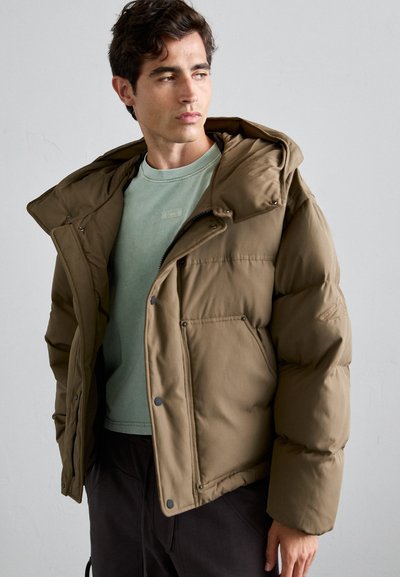 Braune Pufferjacke mit Kapuze, die Druckknopfverschlüsse, Seitentaschen und ein leicht verkürztes Design aufweist. Über einem hellgrünen Hemd getragen.