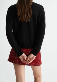 Maglione nero a maniche lunghe abbinato a una gonna in tweed rossa con orlo frangiato e testurizzato. Mani incrociate dietro la schiena.