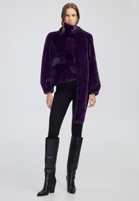 Donna che indossa una giacca in pelo sintetico viola scuro con un lungo dettaglio di sciarpa, pantaloni neri e stivali neri in pelle al ginocchio, in piedi su uno sfondo semplice.