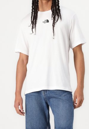 Mand iført hvid The North Face kortærmet t-shirt og blå jeans, vist fra nakke til midt på låret mod ensartet baggrund.
