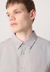 Lichtgrijs linnen shirt met een puntige kraag, een voorplacket en drie knopen. De stof heeft een subtiele textuur en een ontspannen pasvorm.