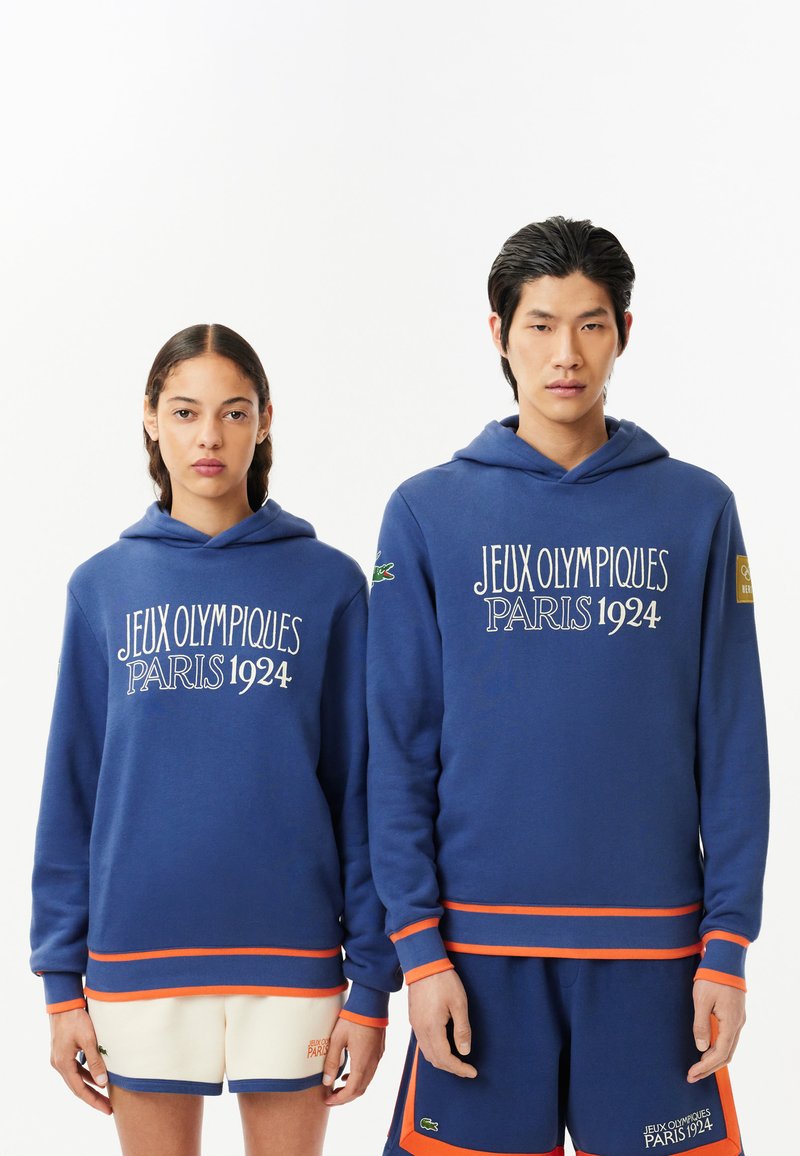 Blaue Hoodies mit weißem Text "JEUX OLYMPIQUES PARIS 1924", versehen mit orangefarbenen Akzenten an den Bündchen und dem Saum, getragen von einem männlichen und einem weiblichen Modell.