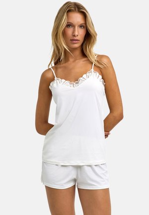 Blonde woman wearing a white sleeveless lace-trim camisole and matching white shorts, standing with hands behind her back.