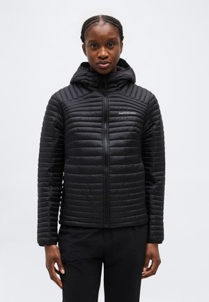 Γυναίκα που φοράει μαύρο κουκουλωτό μπουφάν με φερμουάρ, στέκεται μπροστά σε απλό ανοιχτό γκρι φόντο.