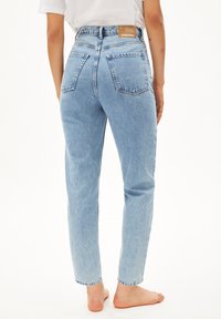 Personne debout pieds nus portant un jean bleu clair taille haute droit avec des poches arrière et un haut blanc.