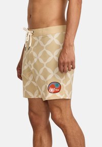 Beige swim shorts with a white geometric pattern, mesh lining, elastic waistband, drawstring closure, and a patch on the left side.